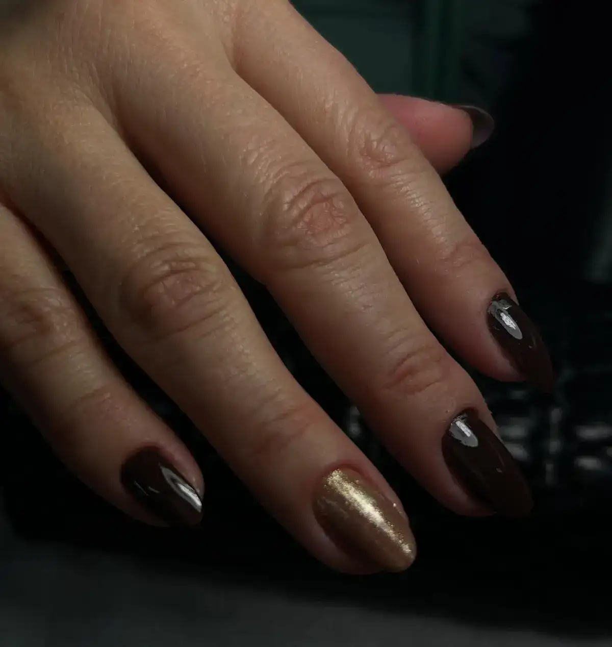 Unhas naturais esmaltadas em marrom intenso, uma com detalhe dourado